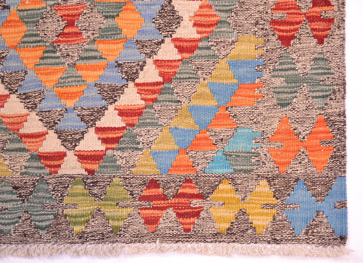 A close-up of a corner of a handwoven small kilim rug showing its geometric pattern with alternating rows of colourful triangles and diamonds in orange, blue, yellow, green, and red on a brown and beige background.