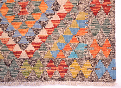 A close-up of a corner of a handwoven small kilim rug showing its geometric pattern with alternating rows of colourful triangles and diamonds in orange, blue, yellow, green, and red on a brown and beige background.