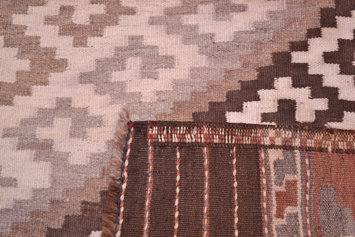 Close-up of the back corner of an old woven Kilim rug showing a geometric pattern in beige, brown, and grey, with fringe and a striped border visible.