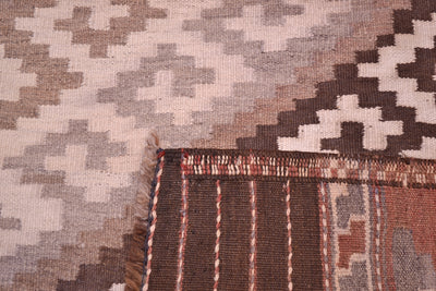 Close-up of the back corner of an old woven Kilim rug showing a geometric pattern in beige, brown, and grey, with fringe and a striped border visible.