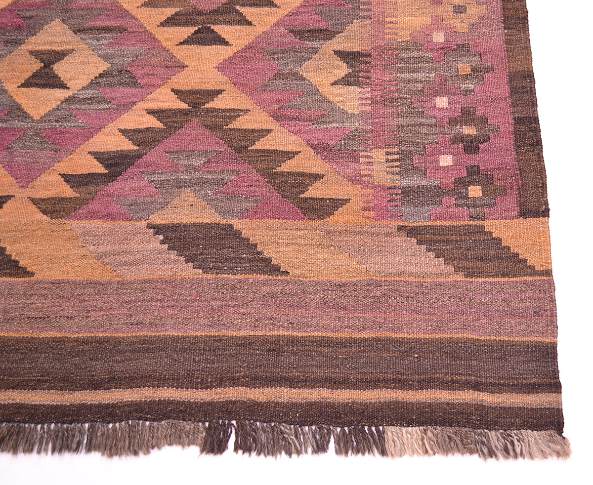 A close-up of a corner of an old handwoven Kilim runner corner showing geometric patterns in dusty pink, brown, tan, and muted grey, with fringe along one edge.