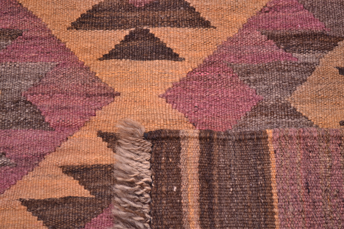 Close-up of the hand woven weave of a geometric Afghan wide Kilim runner with pink, brown, and tan diamond and triangle motifs, showing a fringed edge.