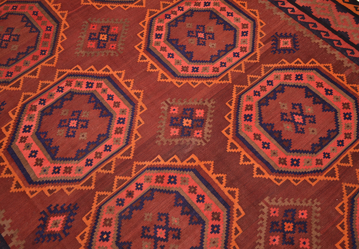A close-up of old handwoven Afghan Kilim rug featuring a repeating geometric pattern with octagonal medallions in rust red, orange, navy blue, and beige.