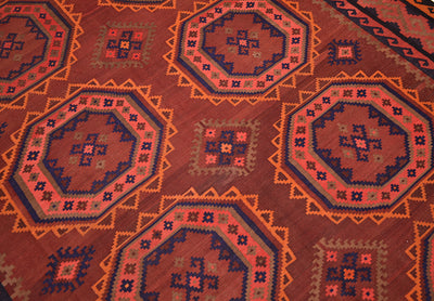 A close-up of old handwoven Afghan Kilim rug featuring a repeating geometric pattern with octagonal medallions in rust red, orange, navy blue, and beige.
