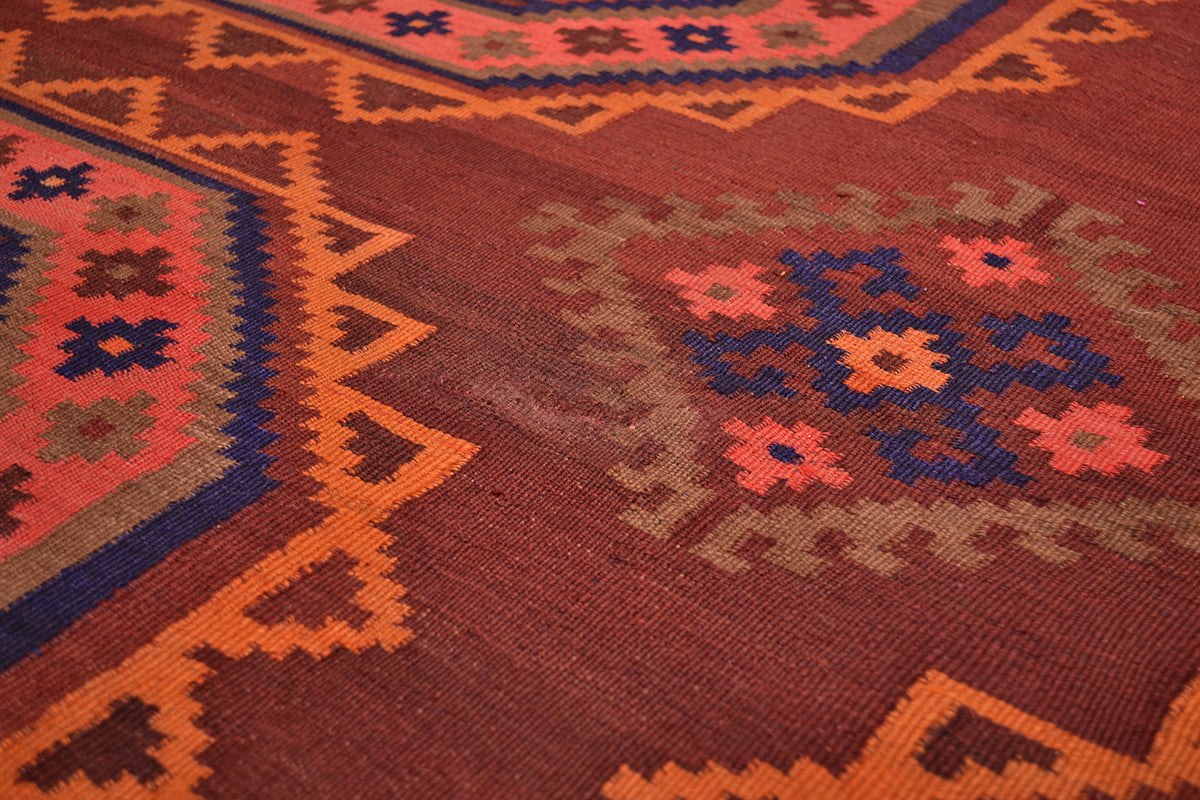 Close-up of a woven kilim rug with a terracotta brown background, featuring geometric borders in orange and blue, and a central floral medallion in coral pink and beige.