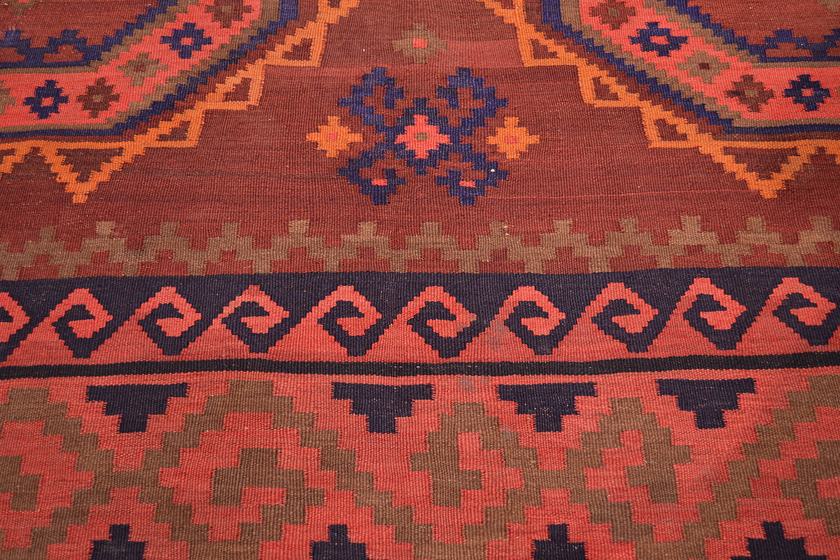 A close-up of a woven kilim rug showing its detailed geometric patterns and color palette, featuring a terracotta red base with navy blue, orange, and olive green motifs.