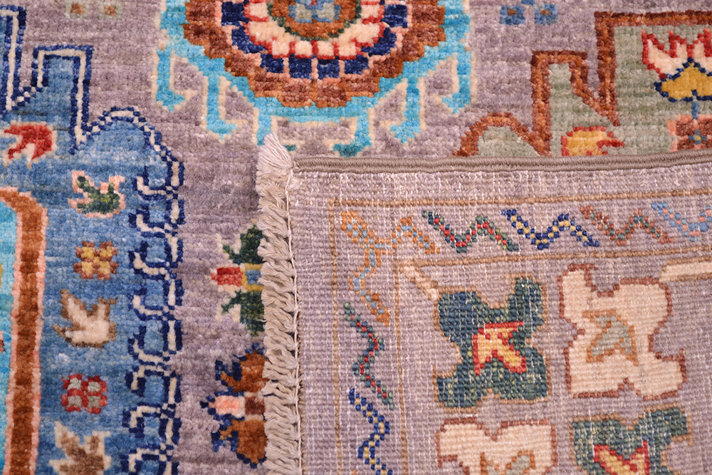 Close-up of the back corner of a hand-knotted Afghan Waziri rug showing a corner with cream fringe, featuring a grey field with blue, brown, green, and beige geometric and floral motifs.