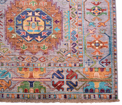 A close-up of a traditional geometric patterned Afghan Waziri rug with a central medallion, border motifs, and fringe at the bottom edge. The design features multiple colours including blue, orange, green, red, and beige on a light greyish field.