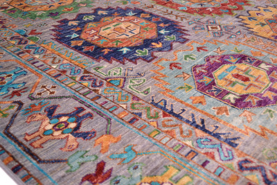A close-up of a colourful patterned Afghan handmade rug with geometric and floral motifs in blue, orange, purple, green, and beige tones on a light grey base.