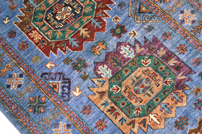 A close-up of a traditional patterned Afghan Wazir runner with a dusty blue field, featuring large central medallions in green, rust red, purple, and tan, surrounded by smaller floral motifs. 