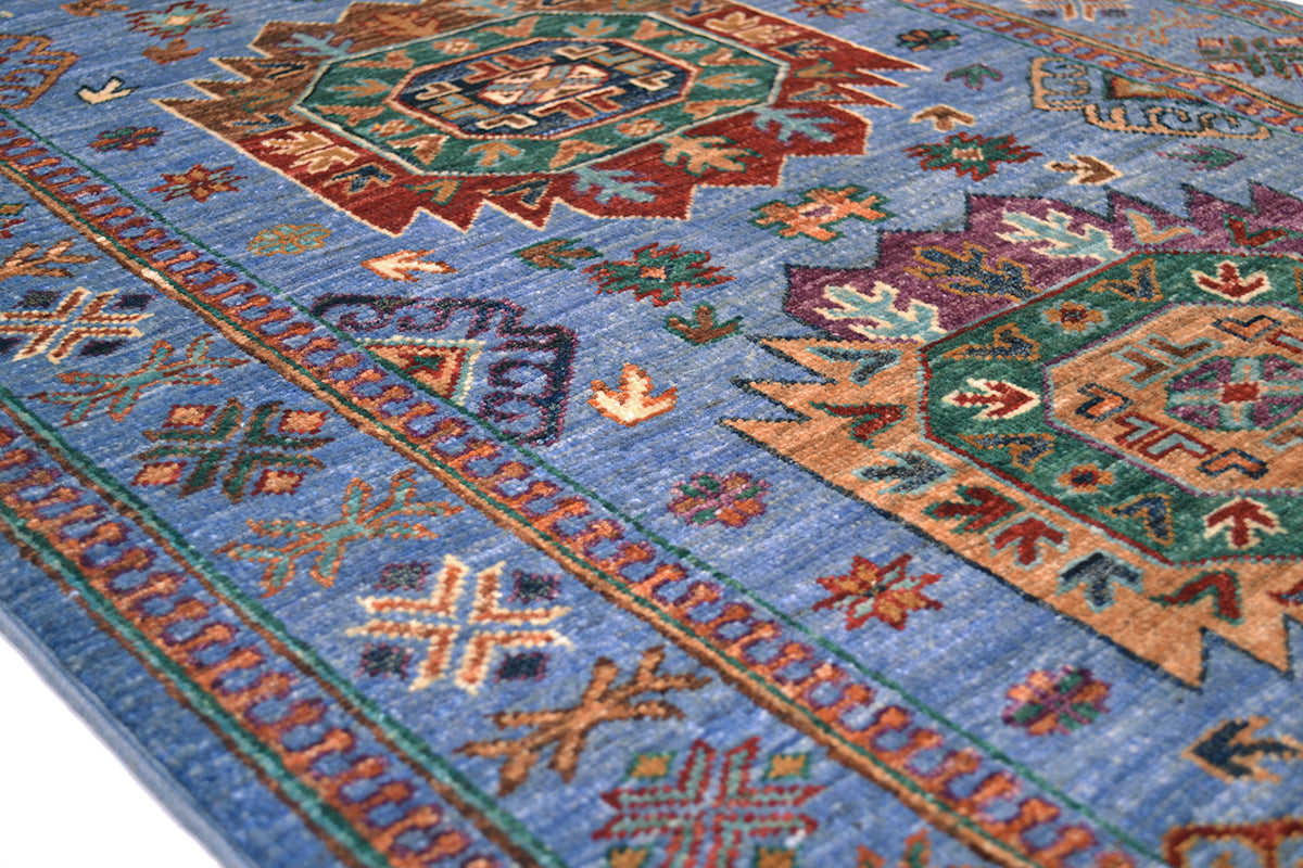 A close-up of a traditional patterned handmade Afghan runner with a medium blue field, featuring geometric medallions and borders in rust red, green, tan, and purple.