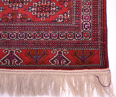 A close-up of a traditional patterned Yamoud rug corner showing a red field with geometric and floral motifs in navy, beige, and orange, finished with a cream fringe.