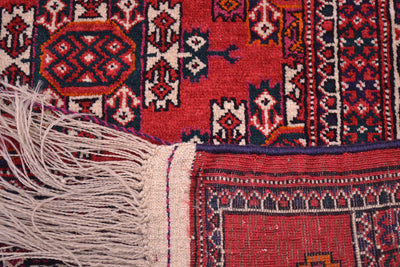 Close-up of corner turned over revealing the hand-knotted weave of a patterned Turkoman Yamoud rug showing a red field with geometric motifs in navy, cream, and orange, plus a cream fringe and border.