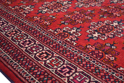 A close-up of a traditional handmade patterned Turkoman Yamut rug with a vibrant red field, detailed geometric motifs, and a dark navy and cream border.