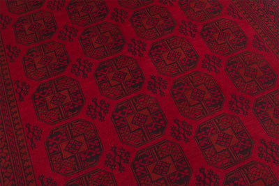 A close-up of a red Afghan Aqcha carpet featuring a repeating dark geometric pattern with octagonal medallions and small floral motifs.