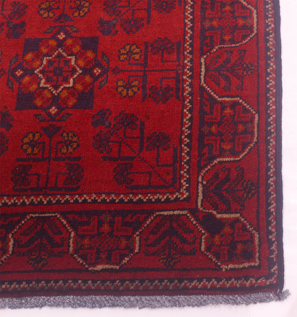 A close-up of a red afghan  rug corner showing a dark navy border, geometric motifs, and a gray fringe.