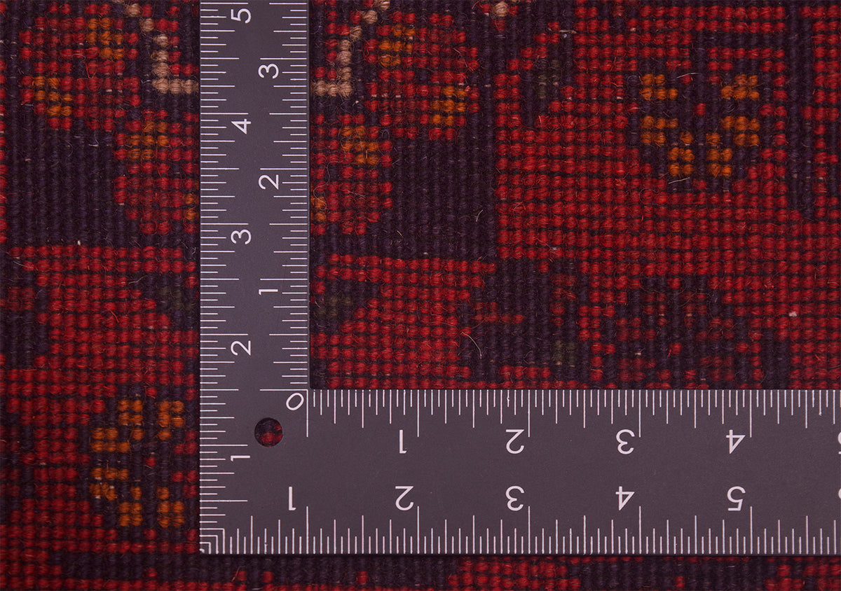 Close-up of a back of a patterned afghan rug with a dark red and black floral design, shown with a metal L-shaped ruler for scale.
