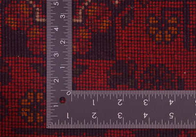 Close-up of a back of a patterned afghan rug with a dark red and black floral design, shown with a metal L-shaped ruler for scale.